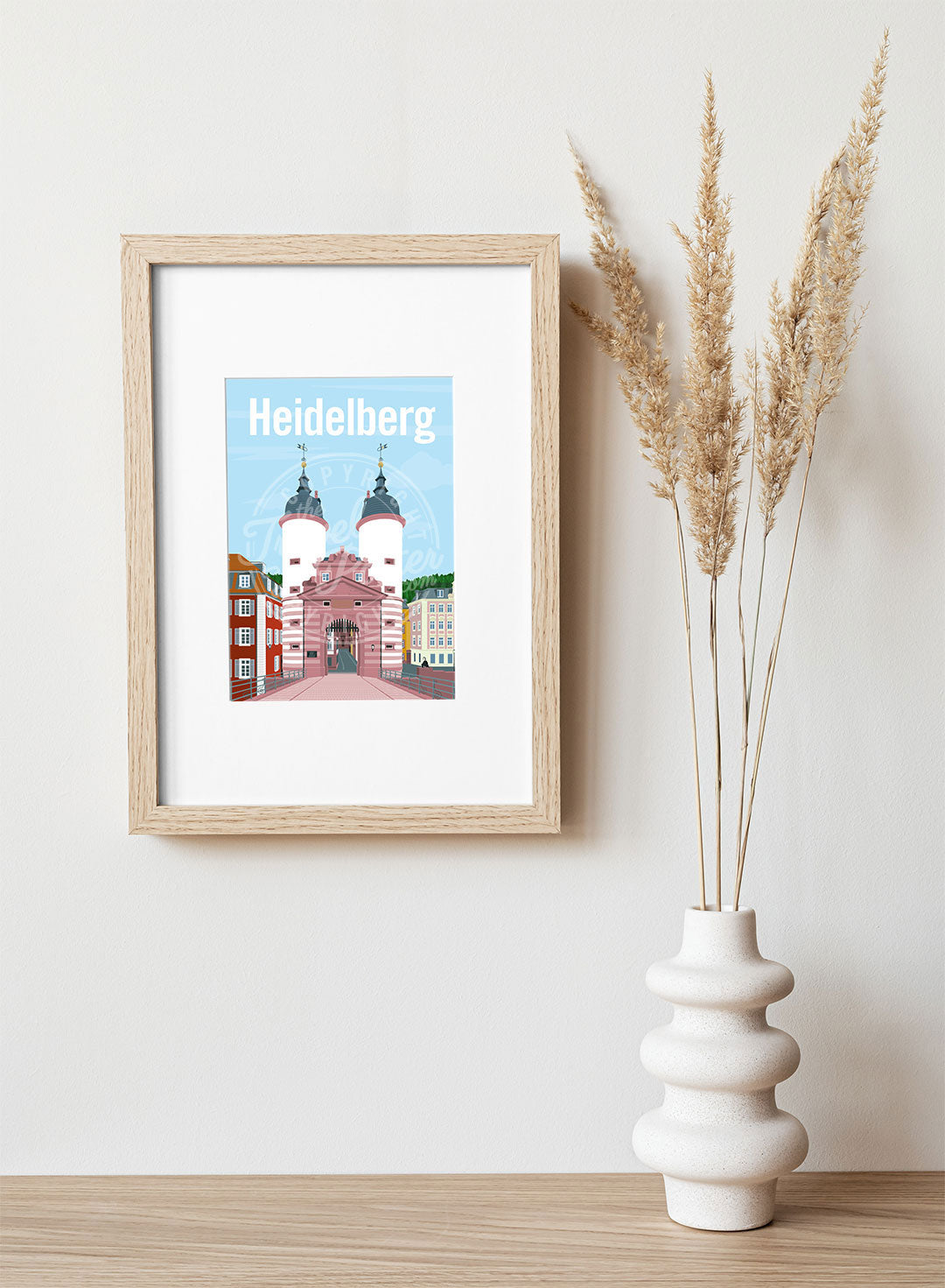 A frame with a postcard of the Heidelberg Bridge Gate, hung on a white wall, above a wooden shelf, on which is a white ceramic vase containing reeds.