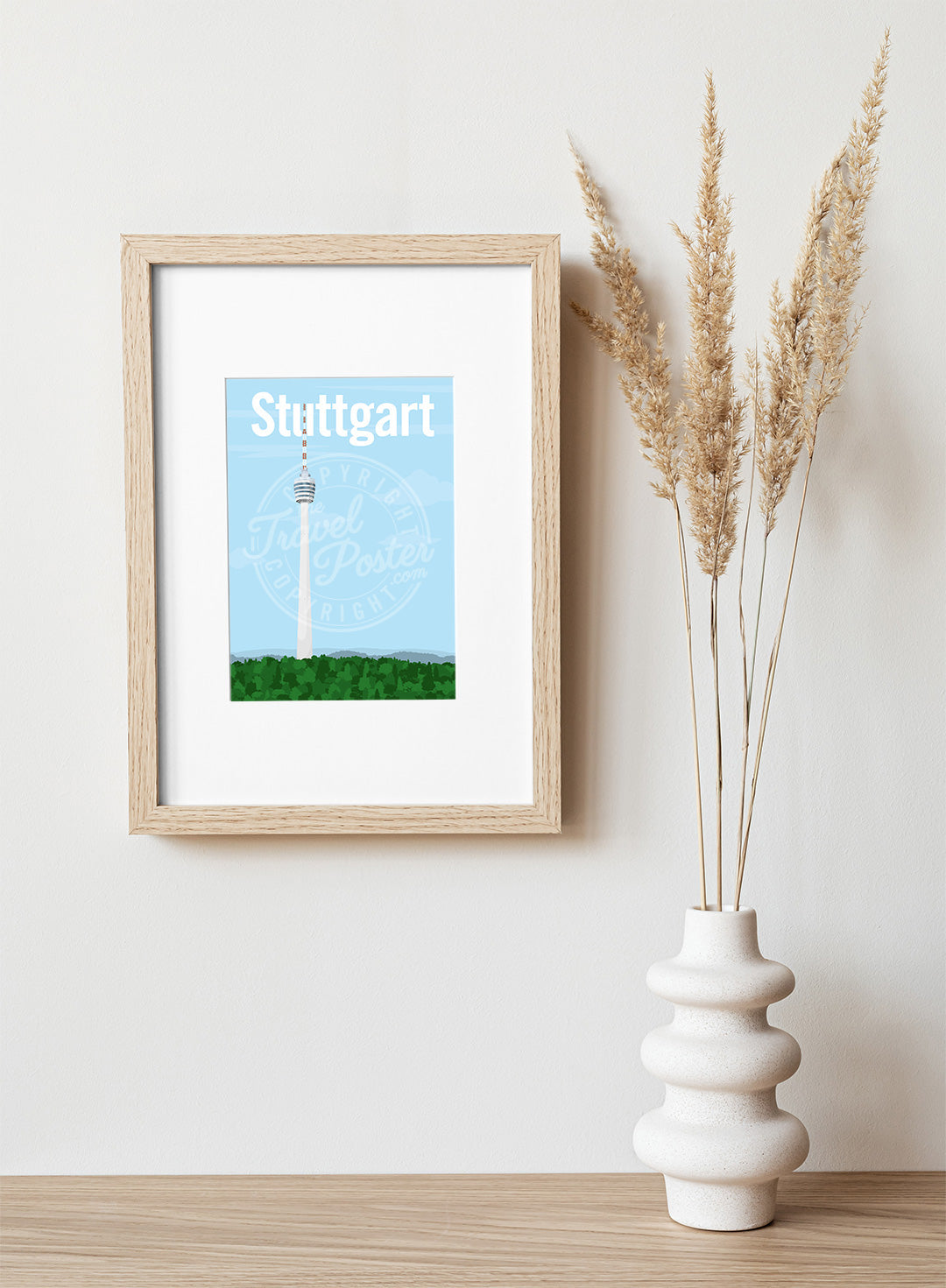 A frame with a postcard of the Stuttgart TV Tower, hung on a white wall, above a wooden shelf, on which is a white ceramic vase containing reeds.