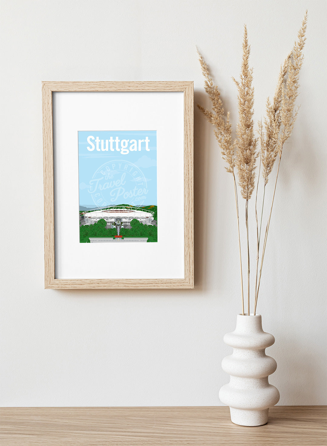 A frame with a postcard of the Stuttgart Neckarstadion, hung on a white wall, above a wooden shelf, on which is a white ceramic vase containing reeds.