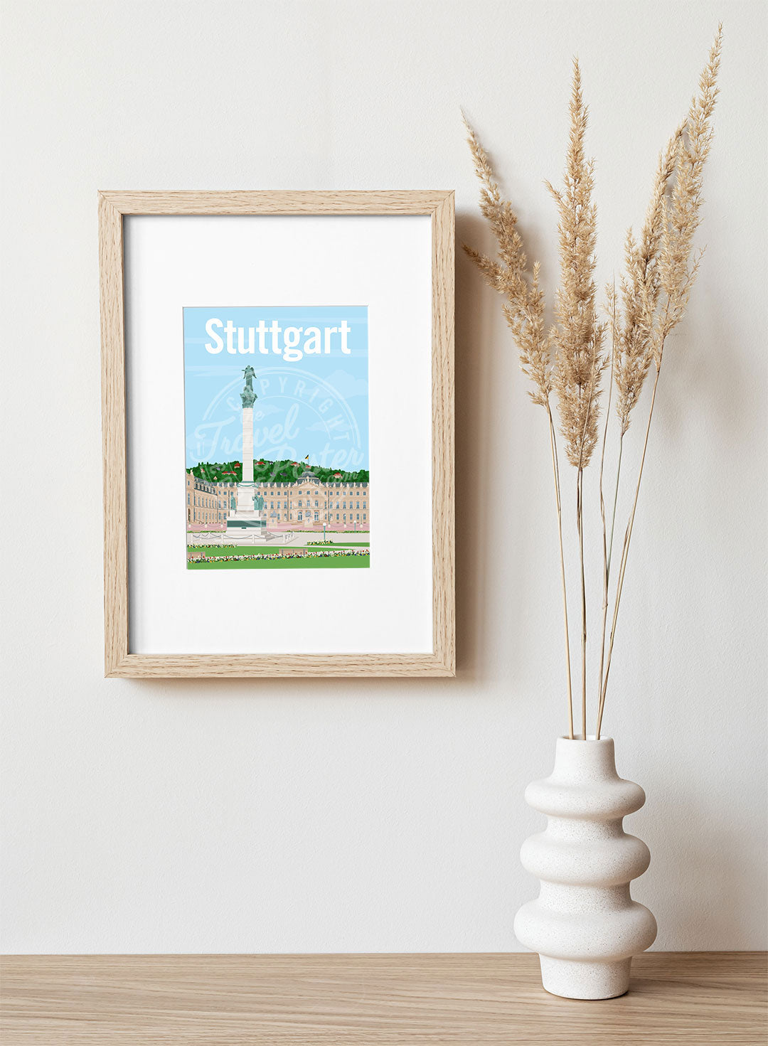 A frame with a postcard of the Stuttgart Schlossplatz, hung on a white wall, above a wooden shelf, on which is a white ceramic vase containing reeds.