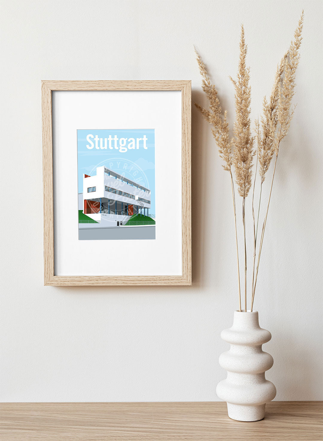 A frame with a postcard of the Stuttgart Weissenhof Museum, hung on a white wall, above a wooden shelf, on which is a white ceramic vase containing reeds.