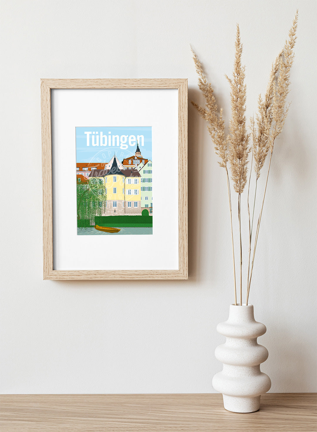 A frame with a postcard of the Tübingen Holderlinturm, hung on a white wall, above a wooden shelf, on which is a white ceramic vase containing reeds.